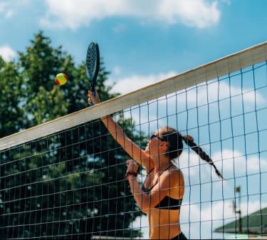 The photo was taken on a beach tennis court. A woman is practicing the modality, jumping above the net height and hitting the ball with a racket.