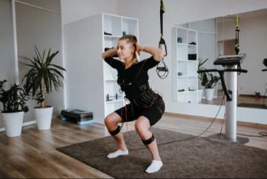 In the photo, there is a woman centered, squatting, stepping on a mat, doing one of the electrostimulation training exercises.