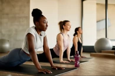 The photo shows three women in a yoga studio. They are in a class, each lying on their own mat, stretching. There is also a ball in the background, which is an accessory used during practice.